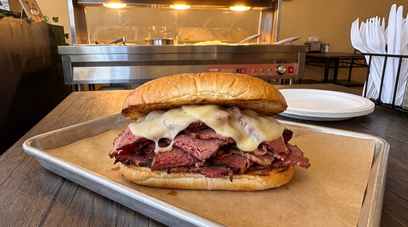 A large roast beef sandwich with melted cheese on a bun, served on a metal tray.